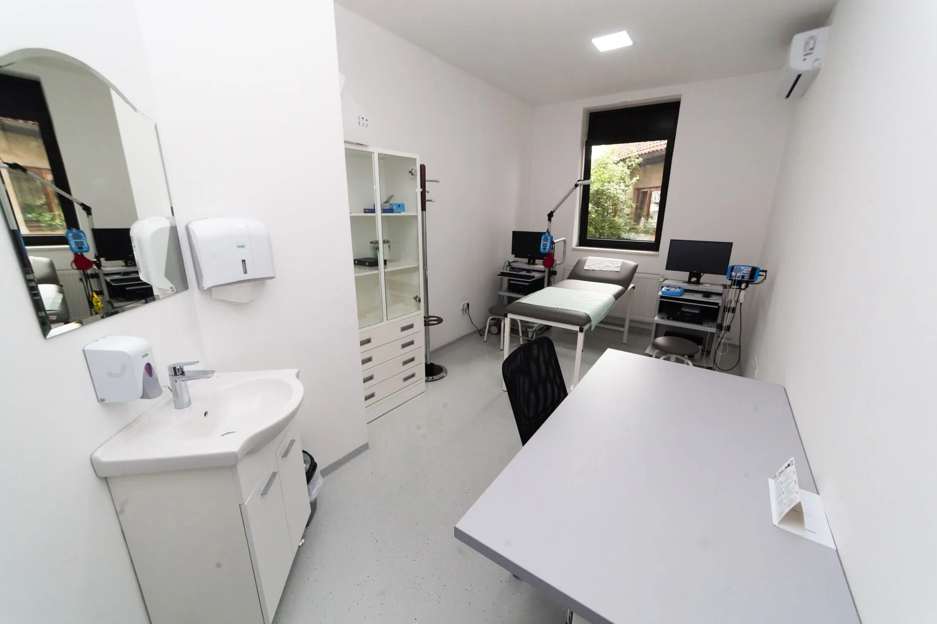A bright clinic room with an examination couch, two workstations and a window onto a leafy courtyard